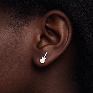 Peut inclure: Une boucle d'oreille en forme de guitare argentée est portée sur une oreille. La boucle d'oreille est un design simple et minimaliste, avec un petit contour détaillé d'une guitare électrique. La boucle d'oreille est placée sur un fond de peau foncée.
