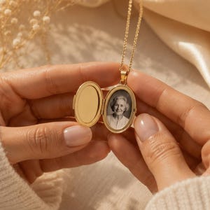 May include: A gold-colored oval locket necklace, open to reveal a black and white photograph. The locket hangs from a delicate chain. The locket is held open by a person wearing a cream-colored sweater.