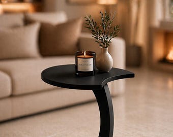 May include: A black, modern side table with a curved leg and a round base. A lit candle in a glass jar and a small vase with greenery sit on the table. The candle has the text "Bibliotheque".