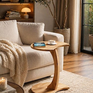 Peut inclure: Une table d'appoint en bois avec un pied incurvé et un plateau en forme de demi-lune. Un livre et une tasse de café sont posés sur la table. La table est à côté d'un canapé beige et d'une fenêtre avec vue sur la verdure.