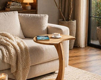 May include: A wooden side table with a curved leg and a half-moon shaped top. A book and a cup of coffee sit on the table. The table is next to a beige sofa and a window with a view of greenery.