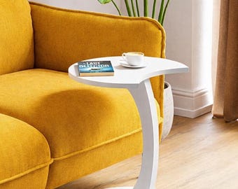 May include: A white, modern side table with a curved design. The table has a rounded base and a half-moon shaped top. A book and a cup of coffee sit on the table. The table is next to a yellow sofa and a large green plant.