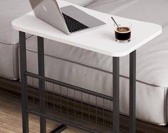 May include: A white laptop table with a black metal frame and a wire mesh magazine rack. A laptop and a glass of coffee with a spoon are on the table. The table is next to a gray sofa.