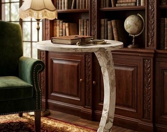 May include: A marble-topped side table with a curved, marble-patterned leg. A book, magnifying glass, and pen rest on the table. The table is in a room with a green armchair, a floor lamp, and a large bookshelf filled with books.