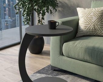 May include: A dark gray C-shaped side table with a circular top and a matching circular base. The table features a curved support column connecting the top and bottom sections. A black mug sits on the flat surface of the table top.
