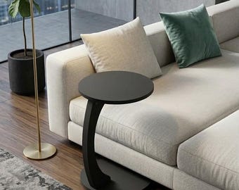 May include: A modern black side table with a circular top and curved support. The table is positioned next to a light-colored sofa with two pillows. A gold floor lamp and a potted plant are also visible.