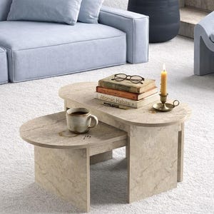 Könnte beinhalten: Ein zweistufiger, beigefarbener Couchtisch aus Stein mit abgerundeten Kanten. Eine Tasse Kaffee, Bücher, eine Kerze und eine Brille liegen auf dem Tisch. Ein hellblaues Sofa und eine Topfpflanze befinden sich im Hintergrund. Der Tisch ist ca. 76 cm lang.