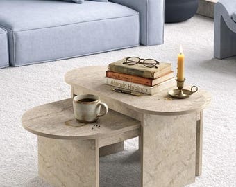 May include: A two-tiered, beige stone coffee table with rounded edges. A cup of coffee, books, a candle, and eyeglasses rest on the table. A light blue sofa and a potted plant are in the background. The table is approximately 30 inches long.