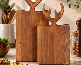 May include: Two rectangular wooden cutting boards with a dark brown finish and a natural wood grain texture. Each board features a handle shaped like a pair of deer antlers at the top. The boards are positioned upright, one slightly behind the other.