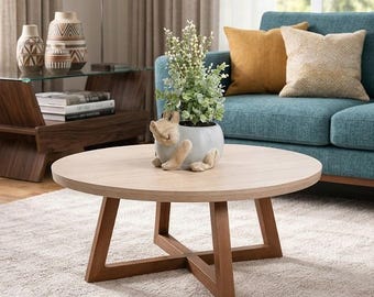 May include: A round coffee table with a light wood grain top and a dark brown wooden cross-base. A light gray ceramic planter shaped like a sitting animal holds a small green plant with white buds, placed in the center of the table surface.