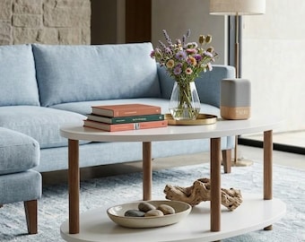 May include: An oval white coffee table with wooden legs and a lower shelf. The top shelf holds books, a vase of flowers, and a speaker. The lower shelf has a decorative bowl with stones and a piece of driftwood. A light blue sofa is in the background.