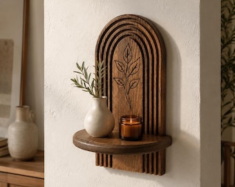 May include: A dark wood wall shelf featuring an arched, tiered design with a carved leaf motif. The shelf holds a small, light-colored ceramic vase containing a green sprig and a glass jar candle with a lit wick, creating a warm, glowing light.