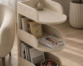 May include: A three-tiered, S-shaped side table with a speckled, off-white stone-like finish. The unit features curved shelves holding books, a magazine, a small beige vase with dried flowers, and a woven basket filled with wrapped candies.