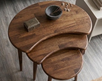May include: A set of three nesting tables crafted from dark brown wood with a crescent-shaped design. The tables feature tapered legs and a smooth finish. A small rectangular wooden box and a grey stone bowl with dried stems sit on the top table.