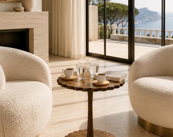 May include: A round wooden side table with a scalloped edge on the top and base, featuring a central pedestal. Two white ceramic cups on wooden saucers, two clear glass tumblers, and a small book are placed on the table surface.