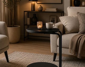 May include: A black metal C-shaped side table featuring a circular top and a matching circular base. The table is equipped with small casters underneath the base for mobility. A glass candle holder with a lit candle and a ceramic mug sit on the top.