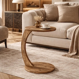 May include: A wooden side table with a curved leg and a round top and base. The table is made of light brown wood and is placed next to a beige sofa. A cup and saucer sit on the table.
