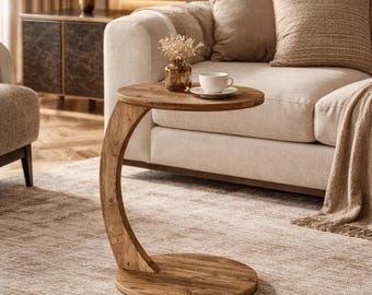 May include: A wooden side table with a curved leg and a round top and base. The table is made of light brown wood and is placed next to a beige sofa. A cup and saucer sit on the table.