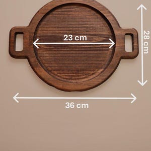 Peut inclure: Un plateau de service rond en bois brun foncé avec deux poignées latérales. Le plateau présente un diamètre intérieur de 23 cm, une hauteur totale de 28 cm et une largeur totale de 36 cm incluant les poignées, selon les flèches de mesure.