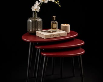 May include: A set of three nesting tables with a deep red finish and black legs. A glass vase with white orchids, a perfume bottle, a candle, and a book titled "KINfolk" are displayed on the tables. The tables are arranged in a descending size.