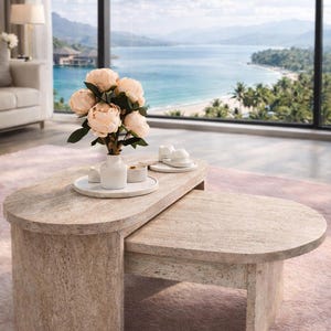 Peut inclure: Un ensemble de tables basses ovales beiges claires. Un vase blanc avec des fleurs p&ecirc;che et de petites assiettes blanches sont pos&eacute;s dessus. La pi&egrave;ce a une grande fen&ecirc;tre avec vue sur une plage et la mer. La grande table mesure environ 120 cm de long.