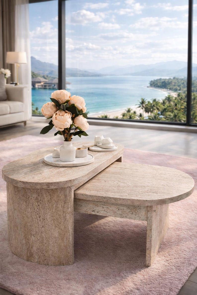 Peut inclure: Un ensemble de tables basses ovales beiges claires. Un vase blanc avec des fleurs p&ecirc;che et de petites assiettes blanches sont pos&eacute;s dessus. La pi&egrave;ce a une grande fen&ecirc;tre avec vue sur une plage et la mer. La grande table mesure environ 120 cm de long.