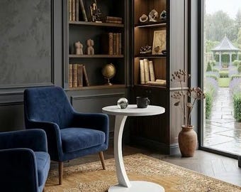 May include: A cozy library setting with two navy blue armchairs, a white side table, and a patterned rug. Bookshelves filled with books and decorative items flank the chairs. A large window offers a view of a garden.