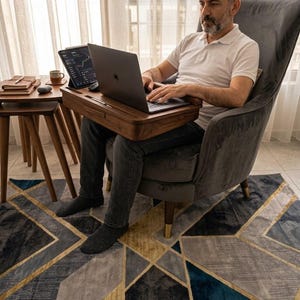 May include: A person seated in a gray armchair using a laptop and tablet on a wooden lap desk. The desk has a built-in slot for the tablet. A small side table holds a coffee cup and notebook. A geometric patterned rug is on the floor.