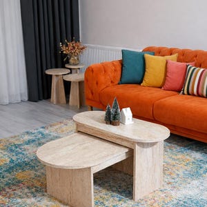 Peut inclure: Salon avec un canap&eacute; orange capitonn&eacute;, des coussins color&eacute;s et un tapis &agrave; motifs. Deux tables basses ovales de couleur claire sont au premier plan, avec de petits arbres d&eacute;coratifs et une maison miniature.