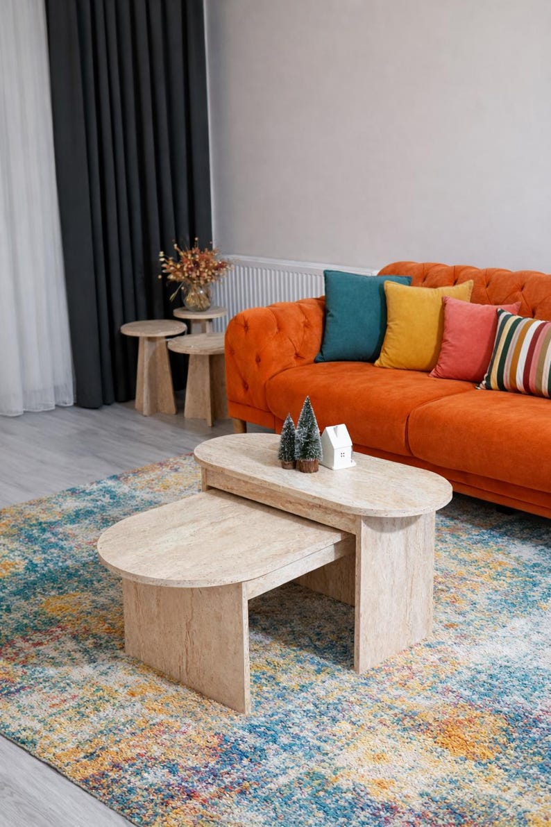 Peut inclure: Salon avec un canap&eacute; orange capitonn&eacute;, des coussins color&eacute;s et un tapis &agrave; motifs. Deux tables basses ovales de couleur claire sont au premier plan, avec de petits arbres d&eacute;coratifs et une maison miniature.