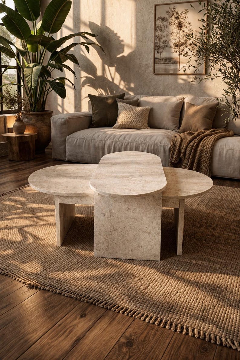 Peut inclure: Une table basse moderne en pierre de couleur claire, avec une surface ovale et deux surfaces circulaires. La table est pos&eacute;e sur un tapis tiss&eacute; dans une pi&egrave;ce avec un canap&eacute; aux tons neutres, des coussins et des plantes. La pi&egrave;ce est baign&eacute;e de lumi&egrave;re naturelle.
