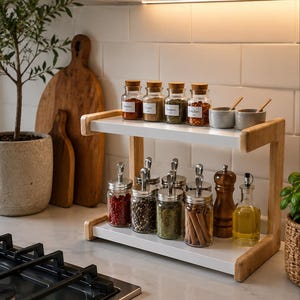 Peut inclure: Une étagère à épices à deux niveaux avec des côtés en bois clair et des étagères blanches. L'étagère supérieure contient quatre bocaux en verre avec des bouchons en liège et deux petits bols gris. L'étagère inférieure contient six bocaux en verre avec des couvercles en métal, un moulin à poivre en bois et une burette à huile.