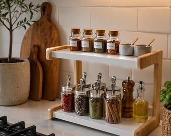May include: A two-tier spice rack with light wood sides and white shelves. The top shelf holds four glass jars with cork stoppers and two small grey bowls. The bottom shelf holds six glass jars with metal lids, a wooden pepper mill, and an oil cruet.