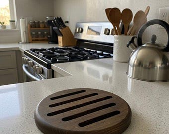 May include: A circular dark brown wooden trivet featuring five parallel horizontal cutouts. The trivet has a smooth, rounded edge and a visible wood grain texture, resting on a light-colored speckled countertop in a kitchen setting.