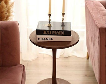 May include: A round dark brown wooden pedestal side table featuring a single central column base. Two tall cream-colored taper candles in metallic holders sit atop two stacked books with black and white covers labeled Balmain and Chanel.