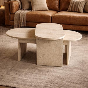 Peut inclure: Une table basse en deux parties de couleur claire, avec un dessus ovale et semi-circulaire. La table est faite d'un mat&eacute;riau semblable &agrave; de la pierre et repose sur un tapis de couleur neutre. Un canap&eacute; en cuir marron et des coussins sont en arri&egrave;re-plan.