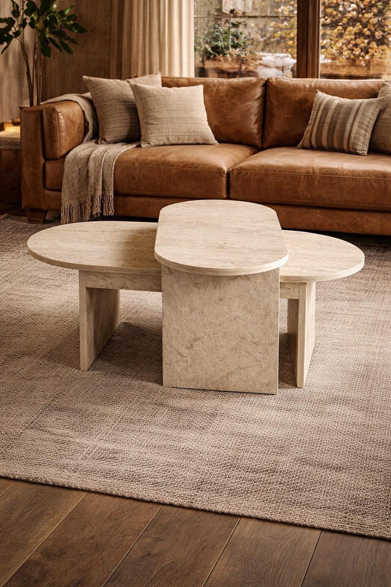 Peut inclure: Une table basse en deux parties de couleur claire, avec un dessus ovale et semi-circulaire. La table est faite d'un mat&eacute;riau semblable &agrave; de la pierre et repose sur un tapis de couleur neutre. Un canap&eacute; en cuir marron et des coussins sont en arri&egrave;re-plan.