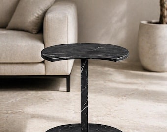 May include: A black marble side table with a semi-circular top and base, connected by a rectangular column. The table has white veining throughout and is set on a light-colored floor. A sofa and potted plant are in the background.