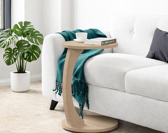 May include: A beige C-shaped side table with a round top and base. A teal throw blanket with fringe is draped over a white sofa. A cup and book rest on the table. A potted plant is in the background.