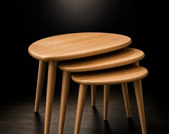 May include: A set of three nesting tables made of light-colored wood. Each table has a rounded oval top and three slender, tapered legs. The tables are arranged in a stacked configuration, with the smallest table at the bottom and the largest at the top.