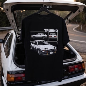 Può includere: Una t-shirt nera a maniche corte con una stampa grafica bianca di un'auto Toyota AE86 Trueno. Il design include il testo TRUENO, una vista laterale, una vista posteriore e una vista frontale di tre quarti dell'auto sportiva bianca e nera.