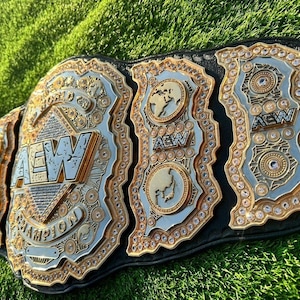 May include: A black leather championship belt featuring large gold and silver metal plates with intricate engravings, embedded clear crystals, and the text AEW World Champion. The central plate displays the AEW logo prominently.