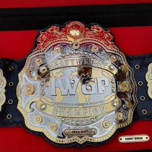 Könnte beinhalten: Ein Wrestling-Meisterschaftsgürtel mit einer großen zentralen Gold- und Silberplatte mit der Aufschrift IWGP HEAVYWEIGHT CHAMPION, flankiert von zwei kleineren Seitenplatten mit eingravierten Namen, auf einem schwarzen Lederriemen.