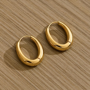 Peut inclure: Une paire de boucles d'oreilles créoles ovales dorées. Les boucles d'oreilles ont une surface lisse et polie et un design simple et élégant. Les créoles mesurent environ 2,5 cm de long et sont posées sur un fond en bois.