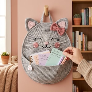 May include: A gray crocheted wall organizer shaped like a cat face with a pink bow, pink inner ears, and pink cheeks. The organizer features a front pocket containing a spiral notebook and a light green to-do list with handwritten text.