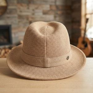 May include: A tan fedora hat with a textured waffle weave design. The hat features a wide brim and a band around the crown. A small decorative emblem is on the band. The hat is sitting on a wooden surface.