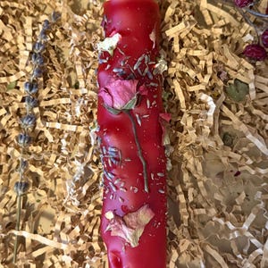 Può includere: Una vivace candela a colonna rossa adornata con petali di rosa secchi e rametti di lavanda. La candela è decorata con fiori ed erbe essiccate. È presentata su un letto di riempitivo di carta marrone, creando un'estetica rustica e naturale.