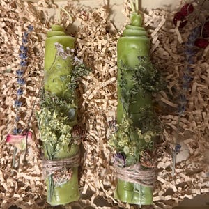 May include: Two green pillar candles adorned with dried flowers and herbs, bound with twine. The candles are presented in a box with shredded paper, lavender and rose hips.