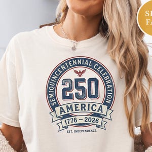 May include: Ivory-colored t-shirt featuring a circular design commemorating the Semiquincentennial Celebration. The design includes the number 250, the word "AMERICA", and the years 1776-2026. The text "EST. INDEPENDENCE" is also present.