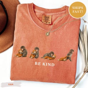 May include: A coral-colored t-shirt with a V-neck and the words "BE KIND" printed on the front. The shirt features an illustration of four monkeys hugging each other. A gold necklace with a pendant is also visible.